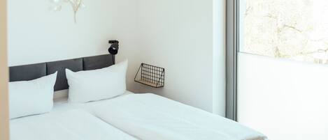 A modern bedroom with a large bed, a small bedside table, a wall-mounted light, and a window with a view of buildings.
