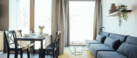 A modern living room with a grey sofa, a dining table with chairs, and a yellow patterned rug.