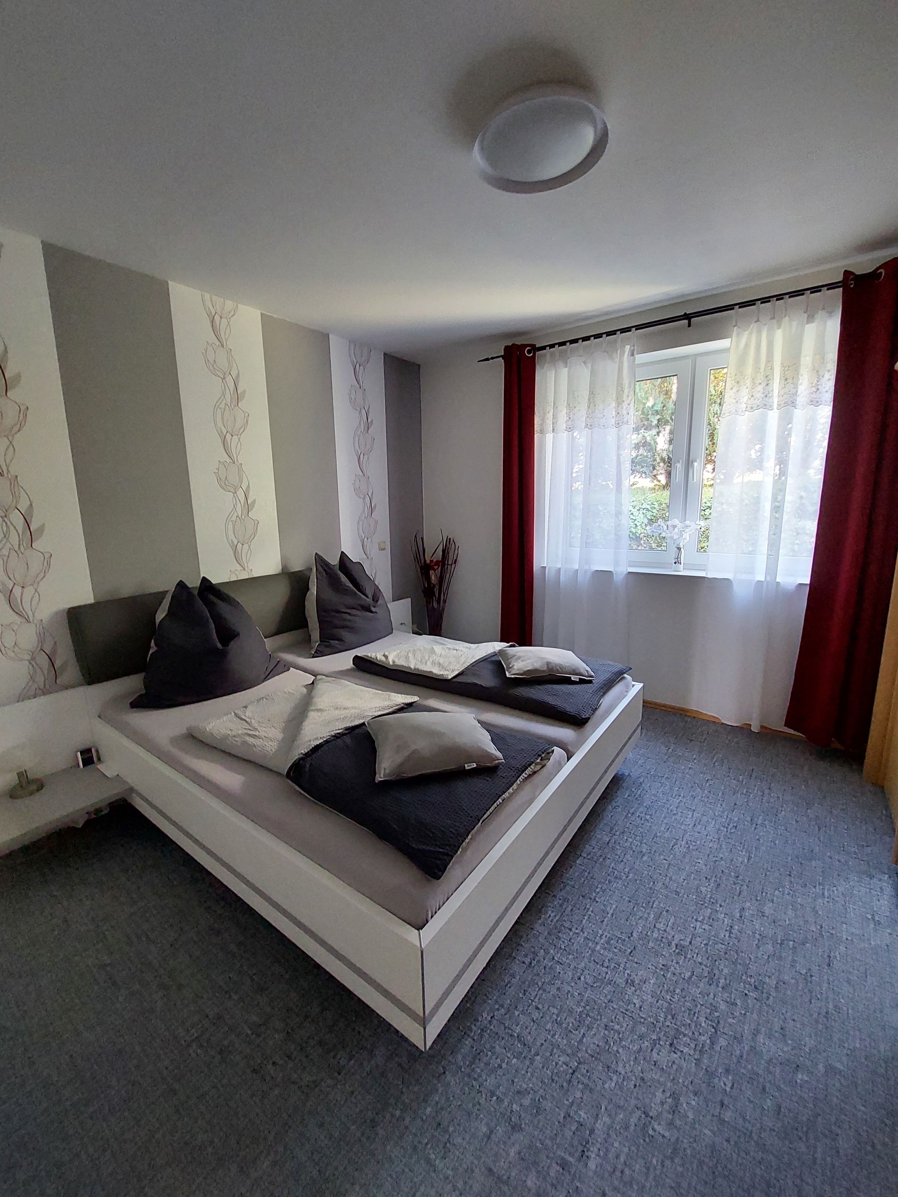 Un dormitorio con cama, mesita de noche, ventana con cortinas y pared con estampado floral.