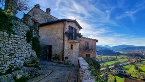 Un edificio de piedra con balcón, un camino adoquinado y vista a un valle con campos y montañas lejanas.
