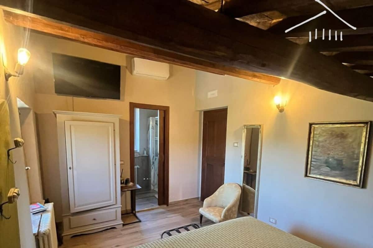 A bedroom with a bed, a chair, a wardrobe, and a mirror. There is a door leading to a bathroom.
