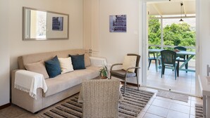 Un salon comprenant un canapé, un fauteuil en osier, une table à manger avec des chaises et une porte-fenêtre coulissante donnant sur un espace extérieur.