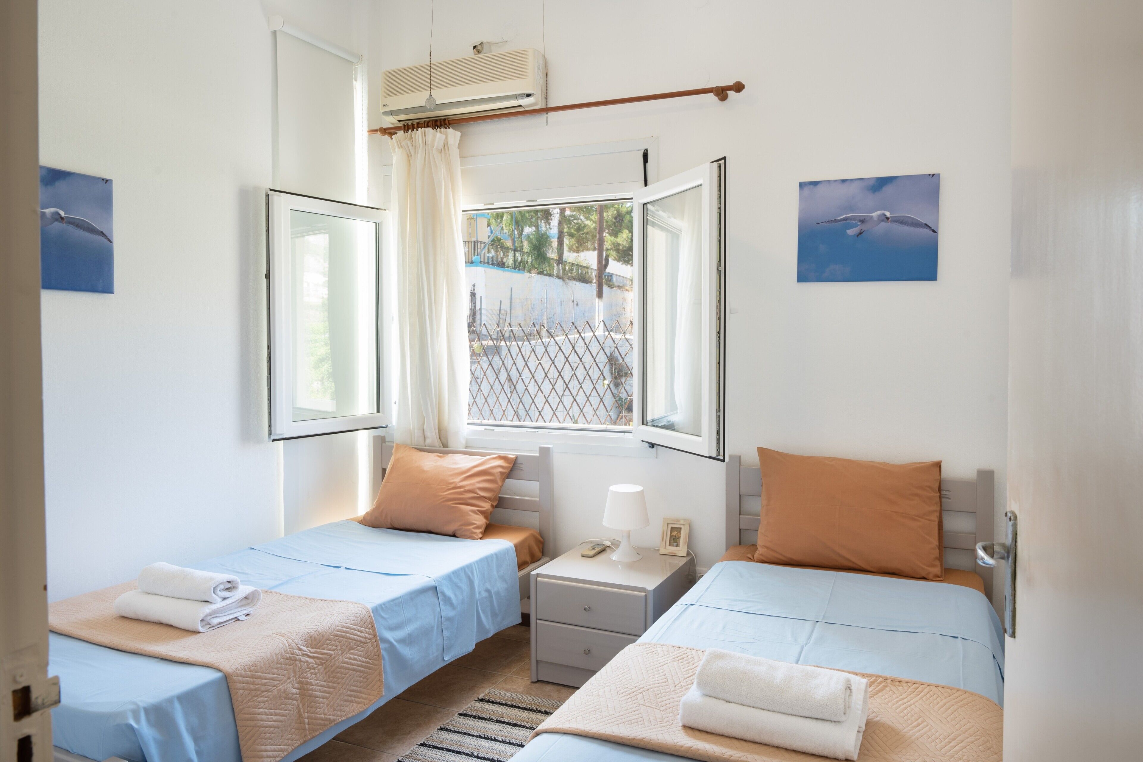 A room with two beds, a window with a view, a bedside table with a lamp, and a wall-mounted air conditioner.