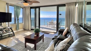 A living room with a leather sofa, a coffee table, a TV, and a balcony with a view of the ocean.