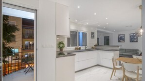 Una cocina moderna con armarios blancos, un comedor con mesa y sillas de madera, y una sala de estar con sofá gris.