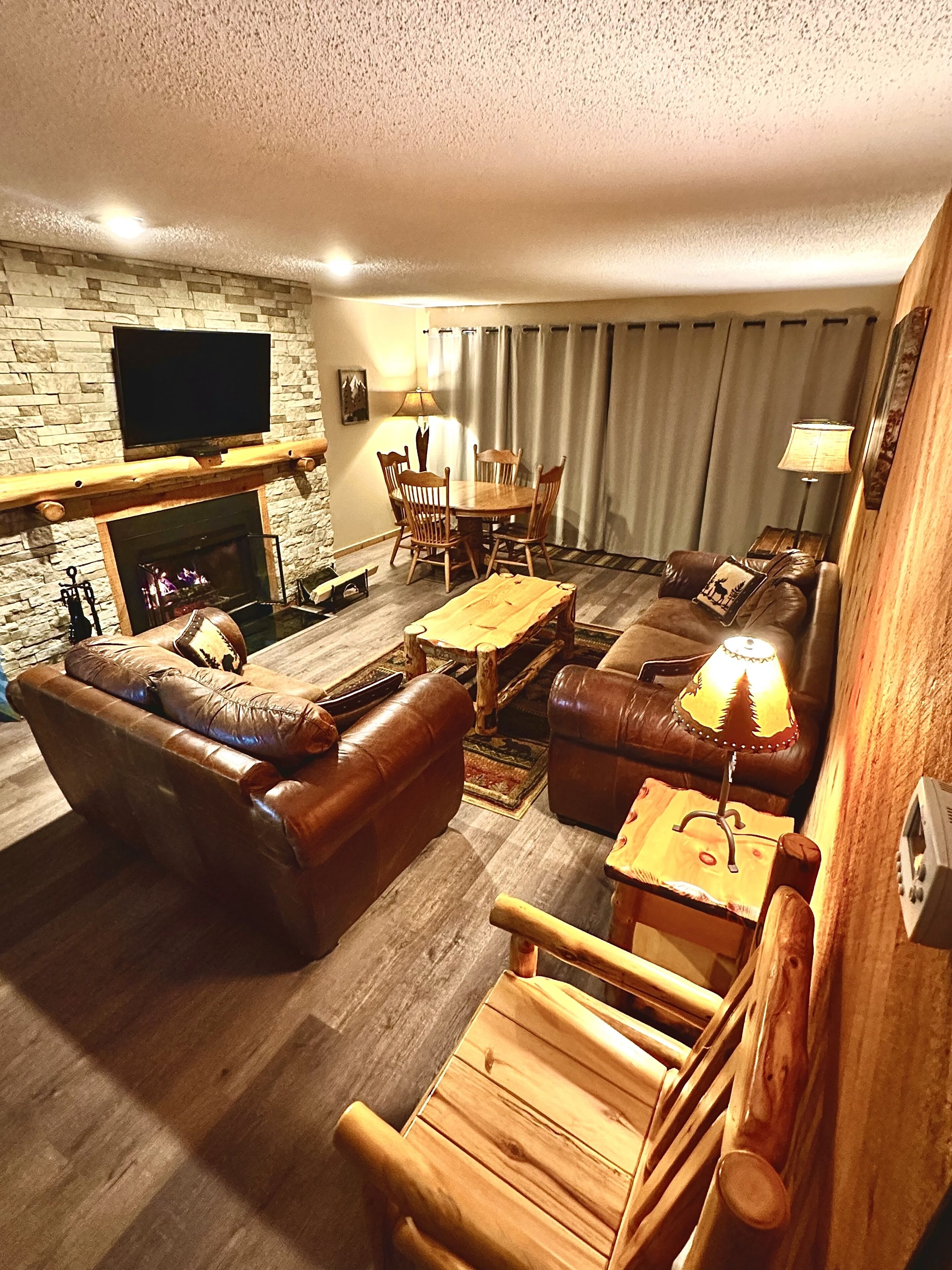 Un salon chaleureux avec une cheminée, des canapés en cuir, une table basse en bois et un téléviseur à écran plat fixé à un mur en pierre.