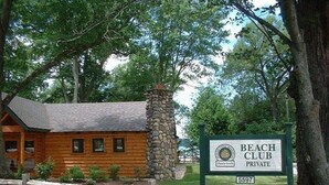 Une cabane en bois avec une cheminée en pierre, entourée d’une végétation luxuriante et d’une clôture en bois. Une pancarte indique qu’il s’agit d’un club de plage privé.