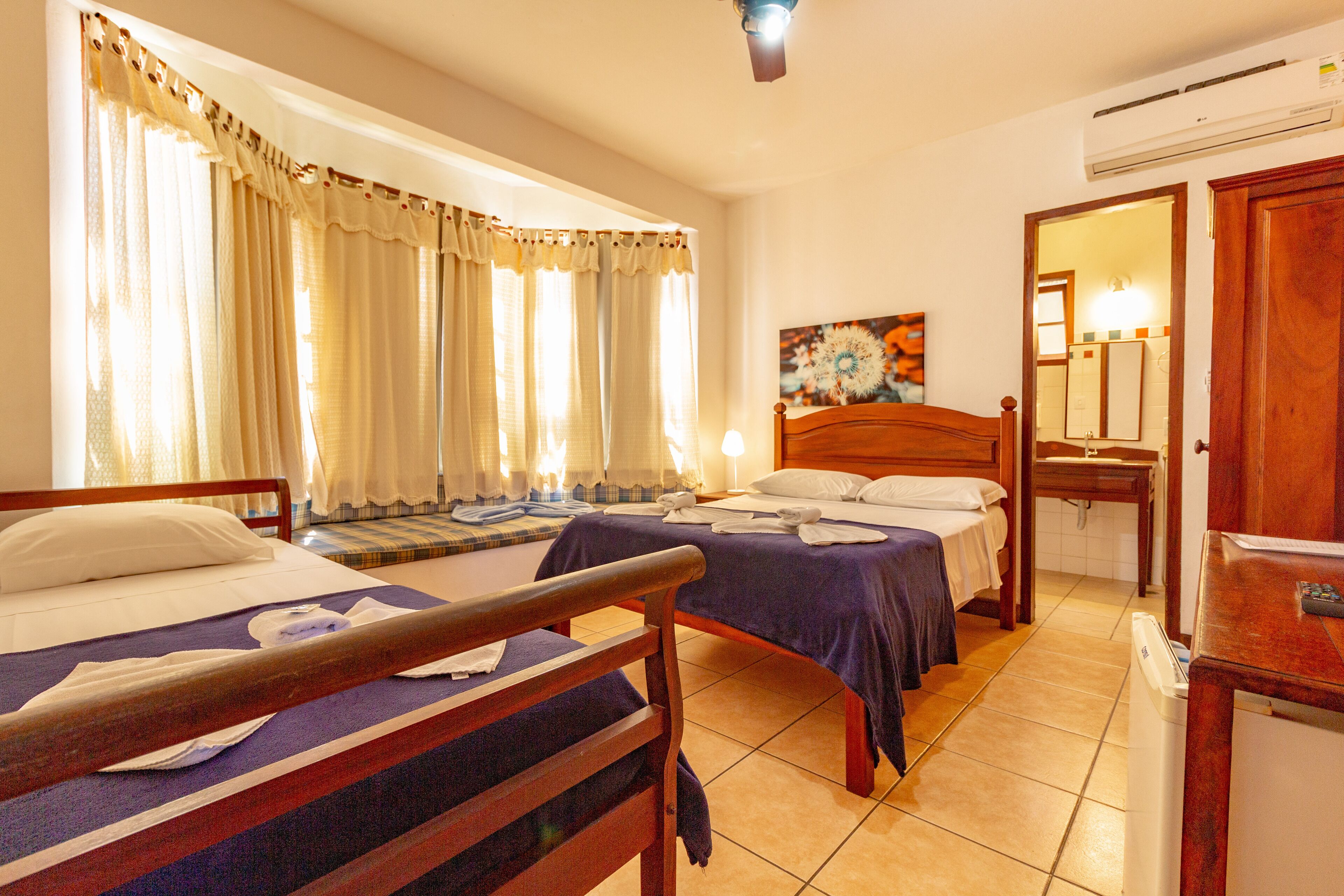Quarto de hotel com duas camas, uma mesinha, um espelho e uma janela com cortinas.