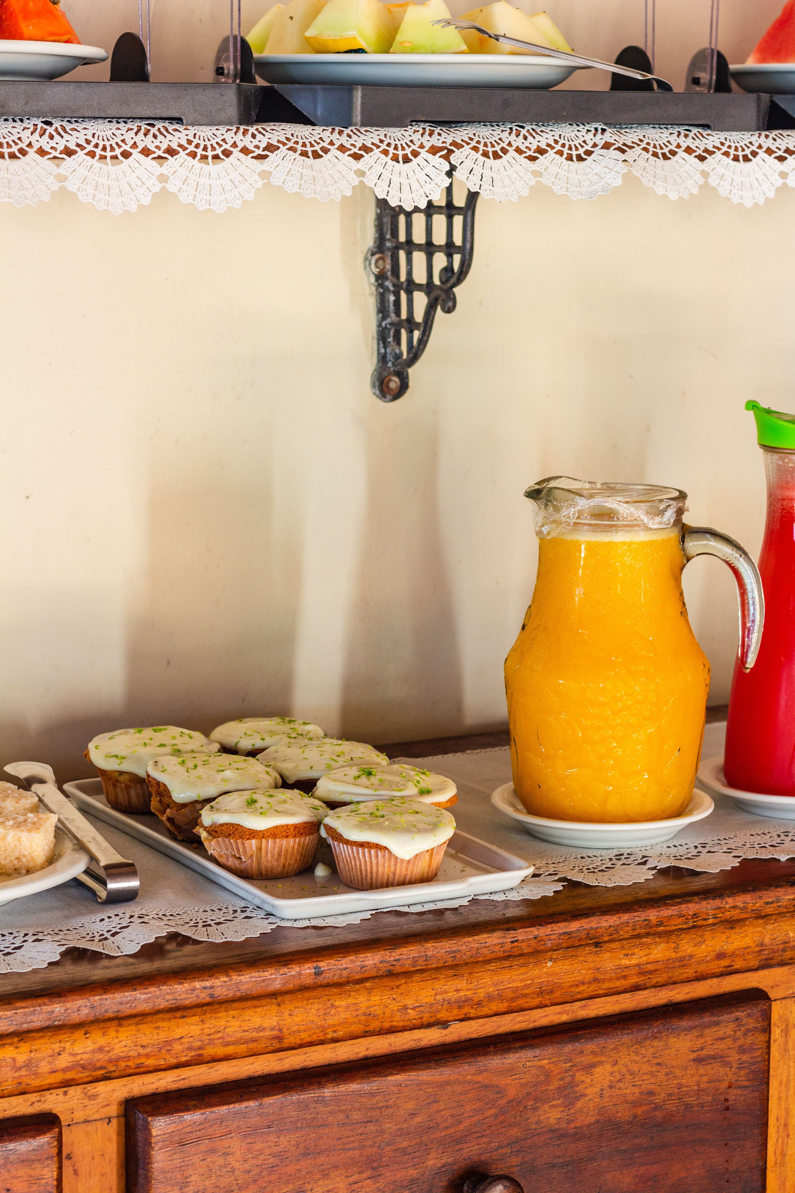 A kitchen counter with a lace doily, a pitcher of orange juice, a red drink, and a tray of cupcakes.