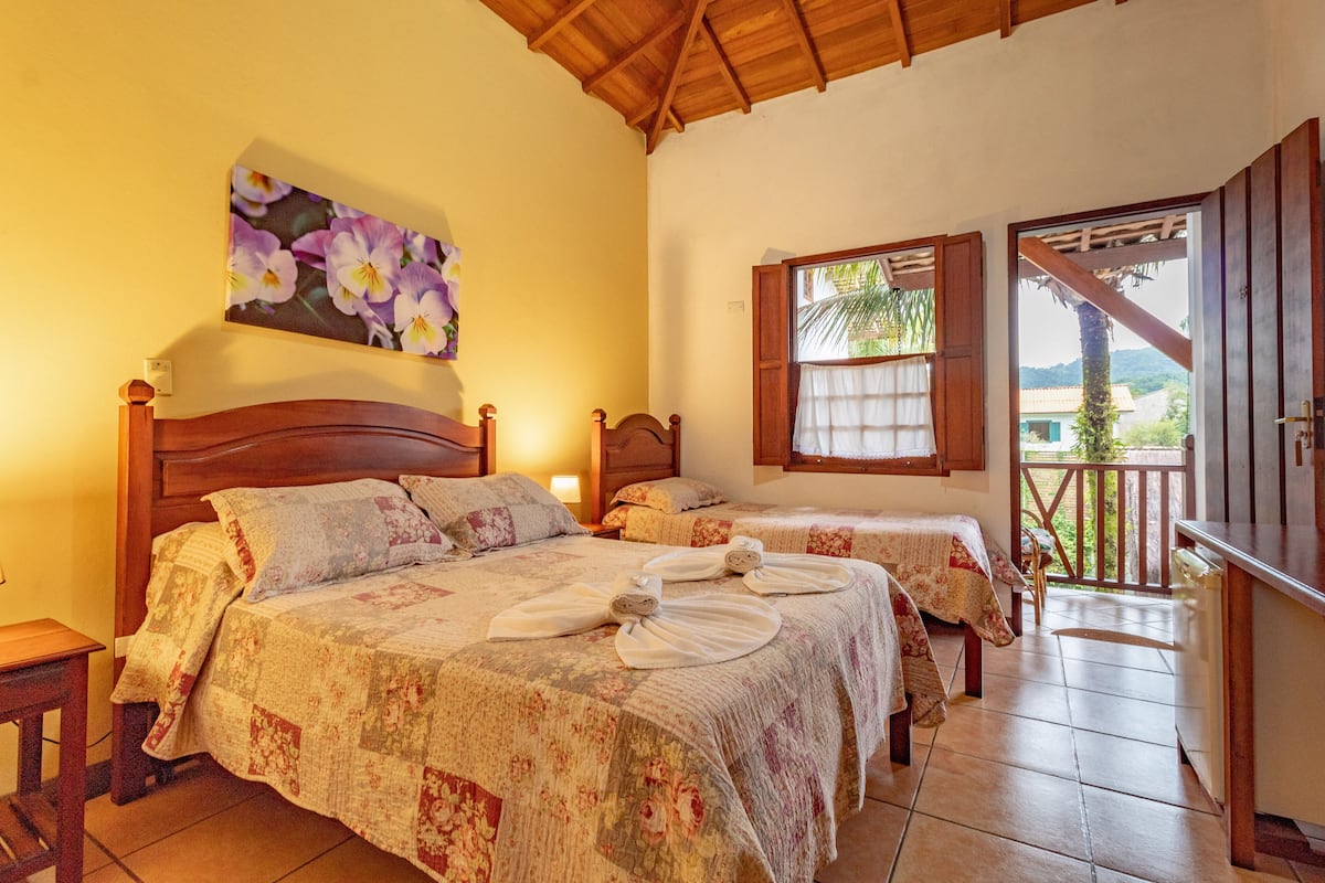 A bedroom with two beds, a wooden ceiling, and a balcony with a view of greenery.