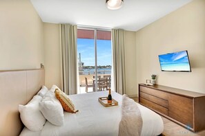 A hotel room with a bed, a TV, a wooden dresser, and a balcony with a view of the sea and a distant building. - Front Bay View Terrace|  Rooftop Pool| Near Beach (Hollywood)
