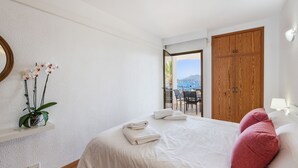 Una camera da letto con un letto, asciugamani bianchi, un cuscino rosso, una pianta in vaso e vista sul mare attraverso una porta aperta.