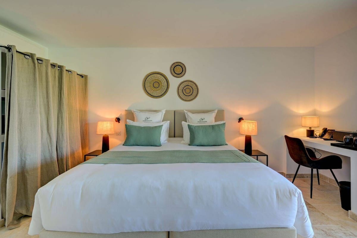 Une chambre d’hôtel avec un lit, deux tables de chevet avec des lampes, un bureau avec une chaise et un rideau sur le côté gauche.