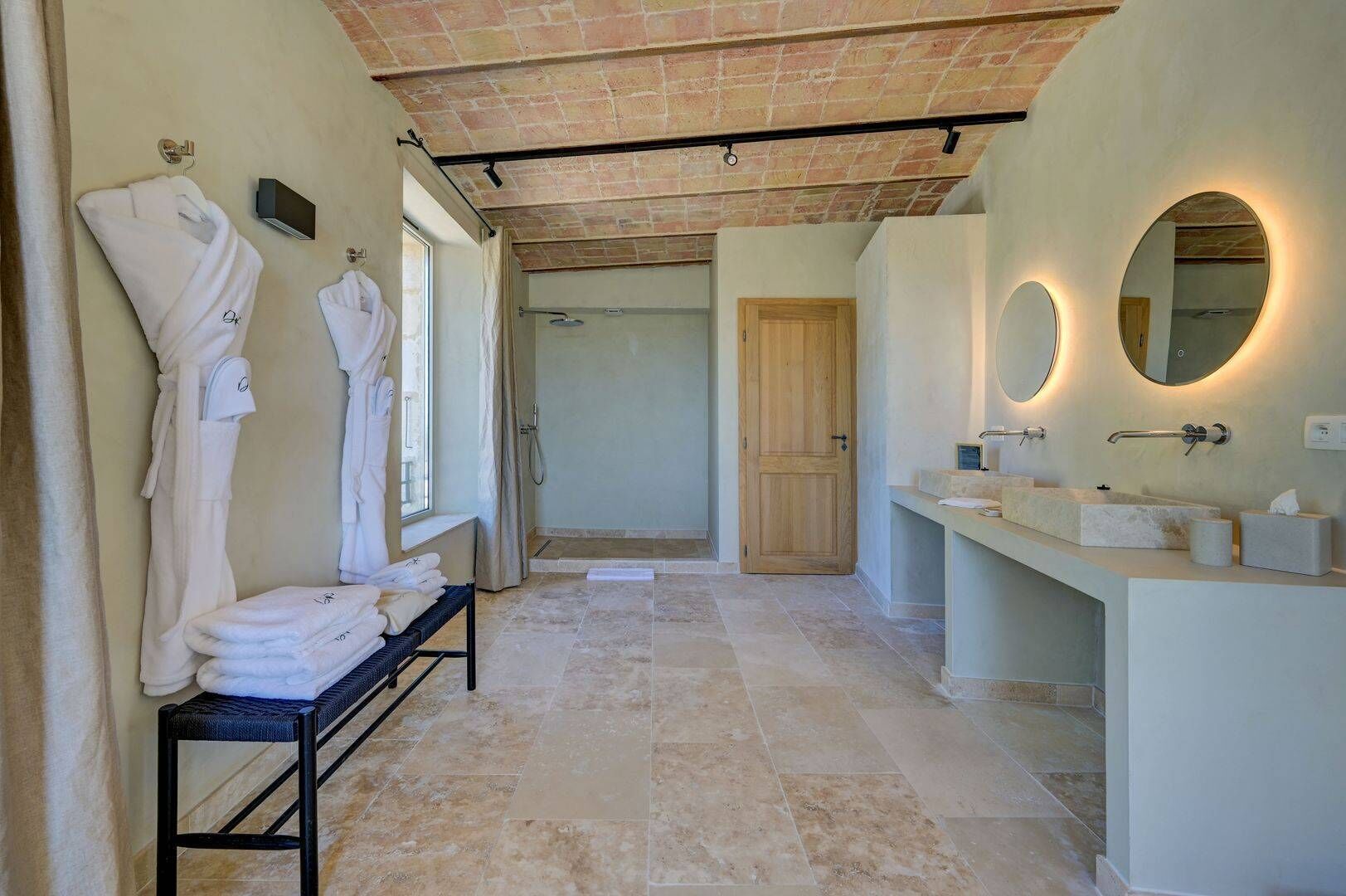 A modern bathroom with a bench holding towels, a wooden door, and a vanity with a mirror.