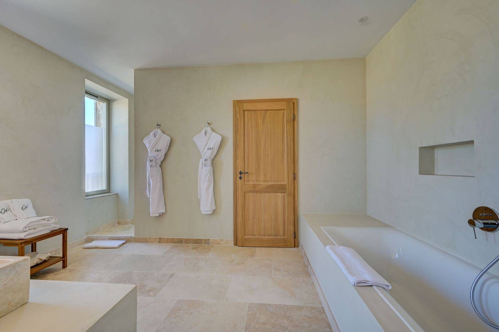 A bathroom with a wooden door, two white towels hanging on the wall, a bathtub, and a small table with folded towels.