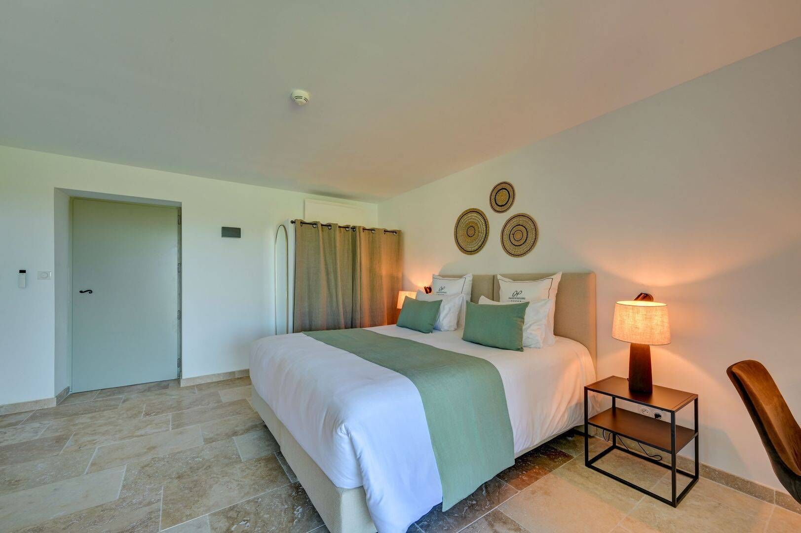 A hotel room with a large bed, a nightstand with a lamp, a chair, and a door.