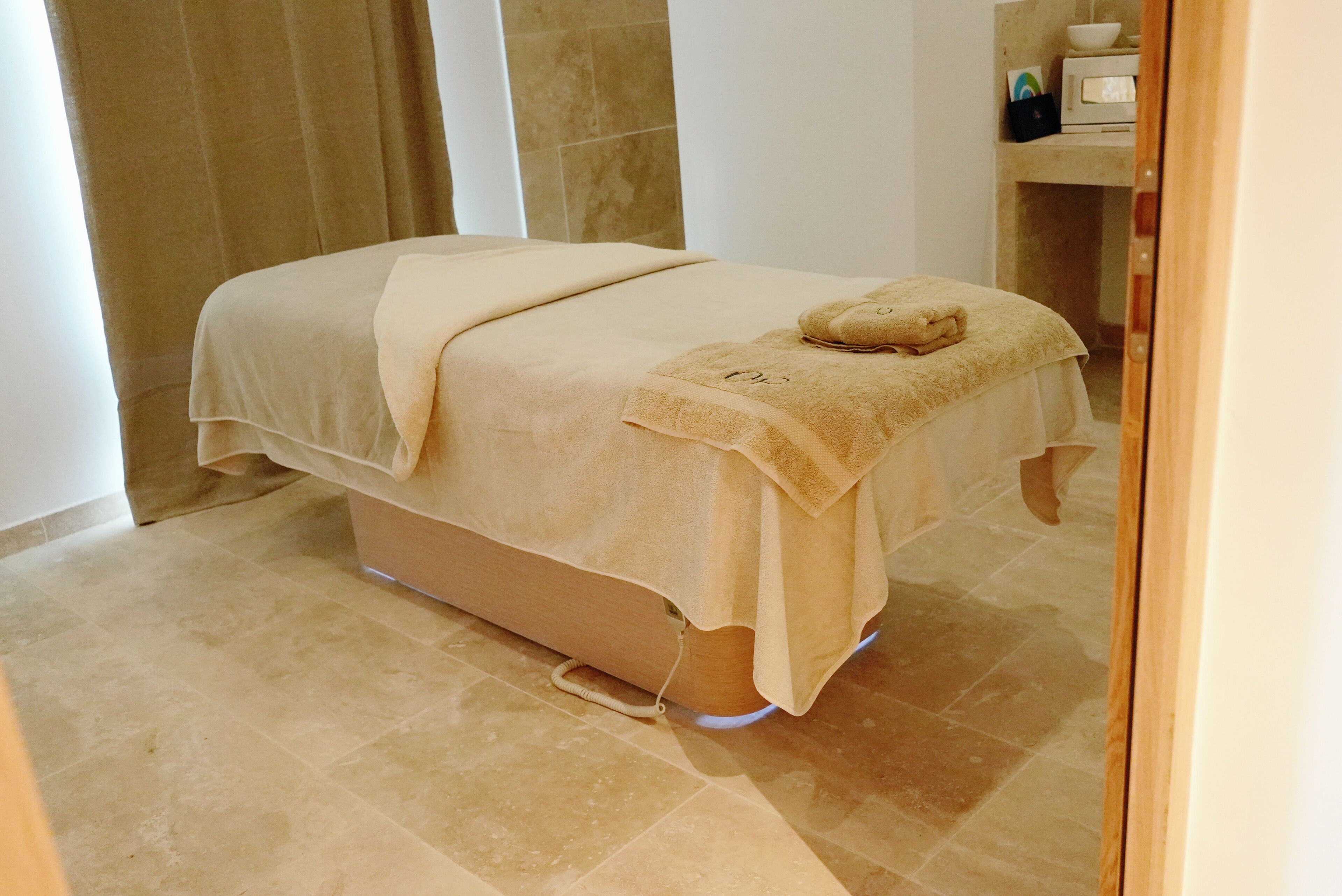A spa bed with a towel, a massage chair, and a small shelf with a bowl and a cup.