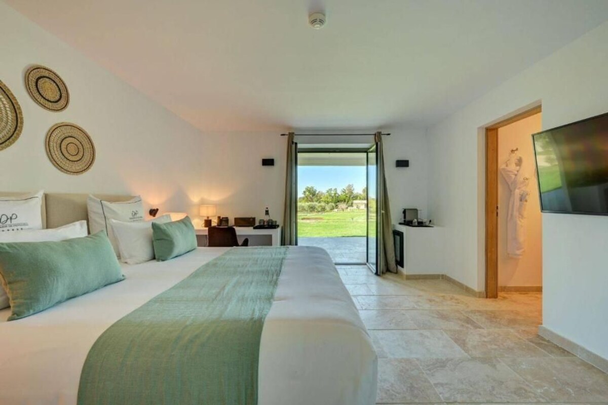 A hotel room with a large bed, a desk, and a TV. There is a view of the outdoors through a glass door.