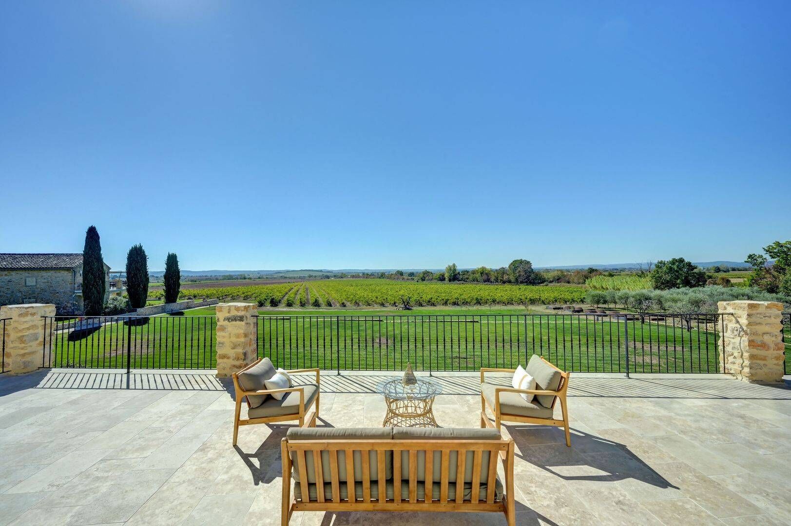 A terrace with two wooden chairs and a small table, overlooking a vineyard and distant hills.