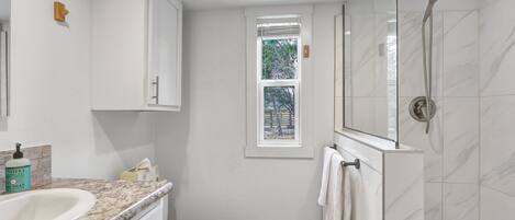 A bathroom with a white sink, toilet, and shower. There is a window with blinds, a towel rack, and a soap dispenser on the counter.