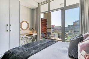 Ein modernes Schlafzimmer mit einem großen Bett, einem Schreibtisch mit Stuhl, einem Spiegel und Blick auf die Stadt durch große Fenster.