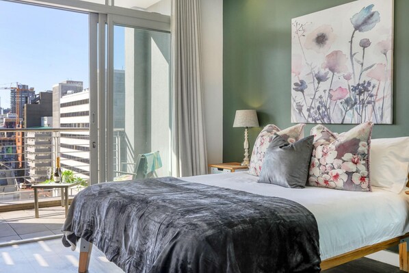 Ein Schlafzimmer mit einem großen Bett, einem Nachttisch, einer Lampe, einem floralen Gemälde und Blick auf die Stadt vom Balkon aus.