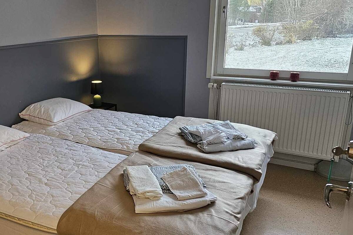 Ein Doppelbett mit zwei Kissen, ein Nachttisch mit Lampe, ein Fenster mit Blick auf eine verschneite Landschaft und eine Heizung.