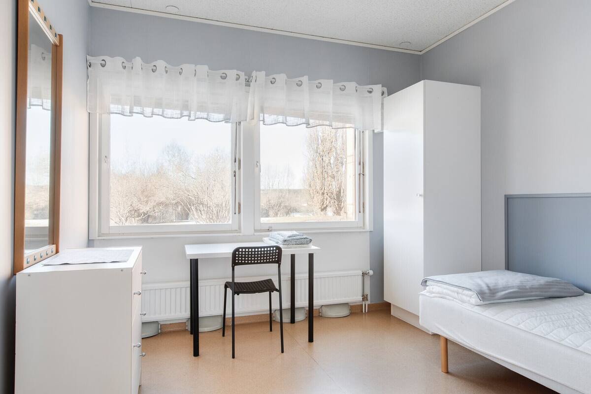 Ein Schlafzimmer mit einem Bett, einem Schreibtisch mit Stuhl, einem Heizkörper und einem Fenster mit Vorhängen.