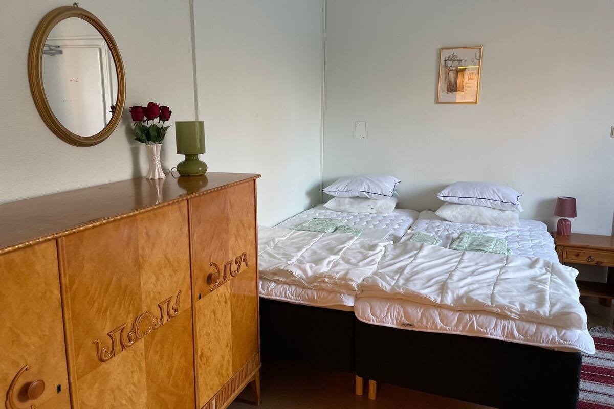 Ein Schlafzimmer mit einem Holzkoffer, einem Bett mit weißer Bettwäsche, einem Spiegel und einem gerahmten Bild an der Wand.
