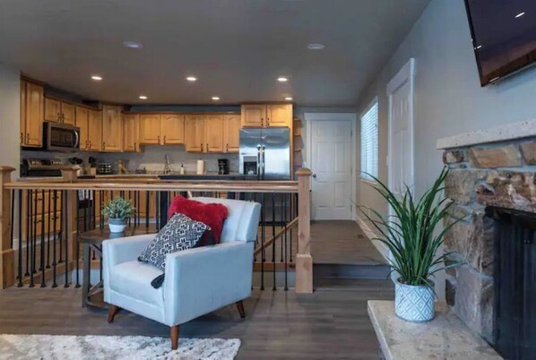 Un salón amplio con un sillón azul claro, una chimenea y una cocina con armarios de madera y electrodomésticos de acero inoxidable.