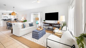 A modern living room with a white sofa, a coffee table, and a TV. There is a kitchen in the background with white cabinets and a dining area.