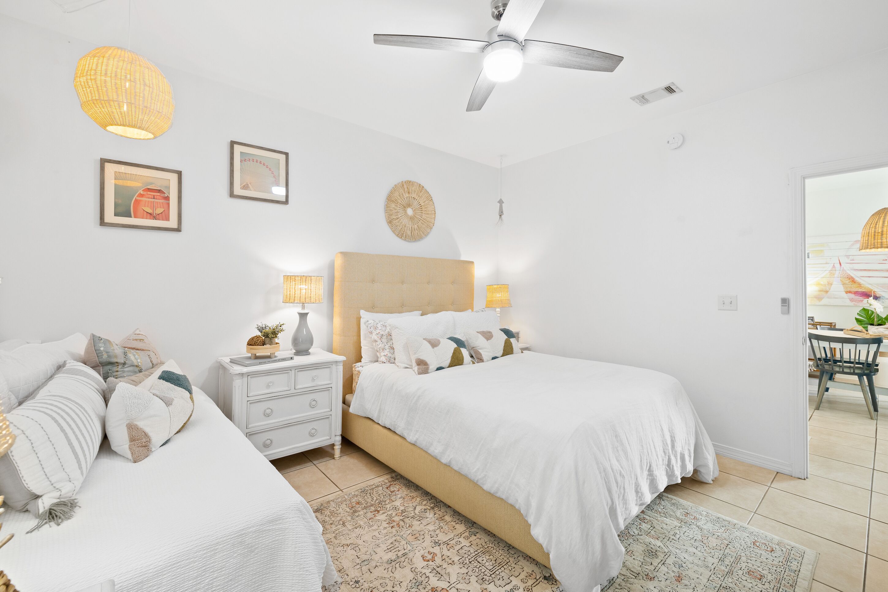 A bedroom with two beds, a ceiling fan, and a dining area visible through an open door.