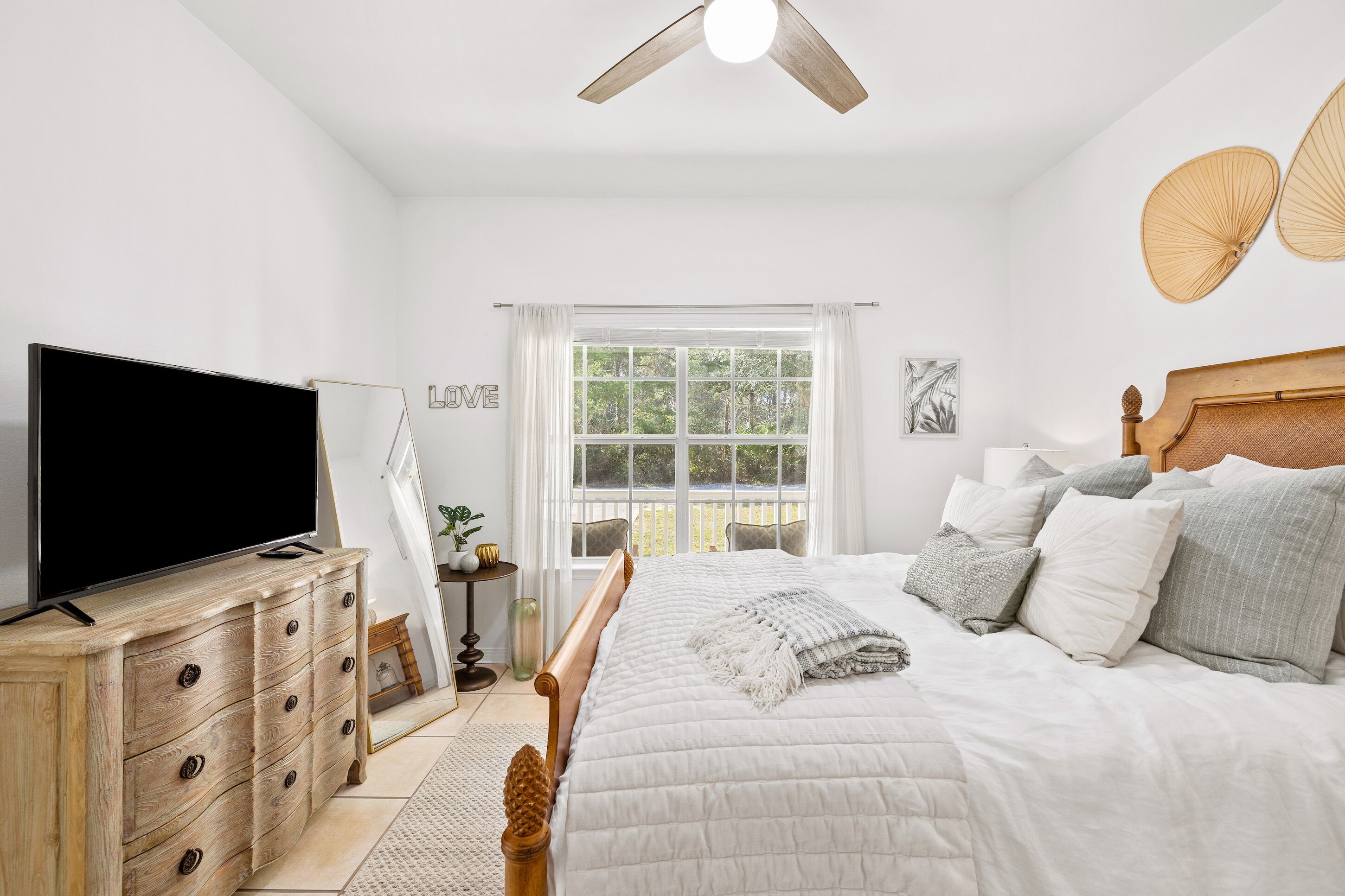 A bedroom with a bed, a TV on a wooden dresser, a ceiling fan, and a window with curtains.
