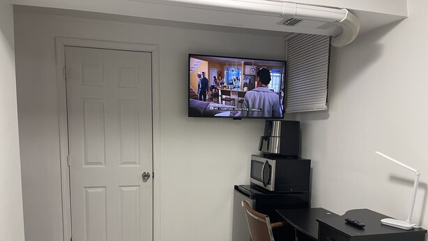 Ett hotellrum med en TV som är monterad på väggen, ett skrivbord med en stol, en mikrovågsugn, ett kylskåp och ett nattduksbord med en lampa.