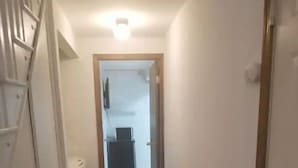 En smal hall med ett mörkt golv, vita väggar och en dörr längst ner som leder till ett rum med ett element och ett litet bord.