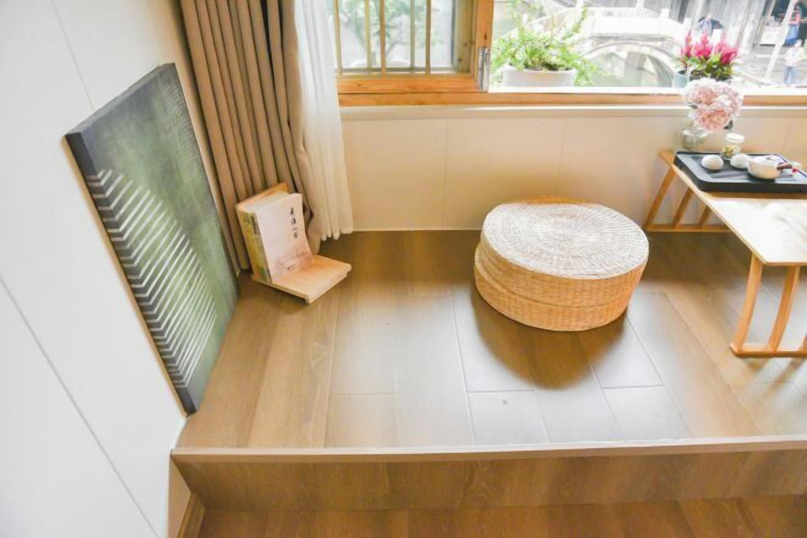 Une pièce avec un plancher en bois, un tabouret rond en osier, un radiateur, une fenêtre avec des rideaux et une petite table sur laquelle est posé un vase de fleurs.