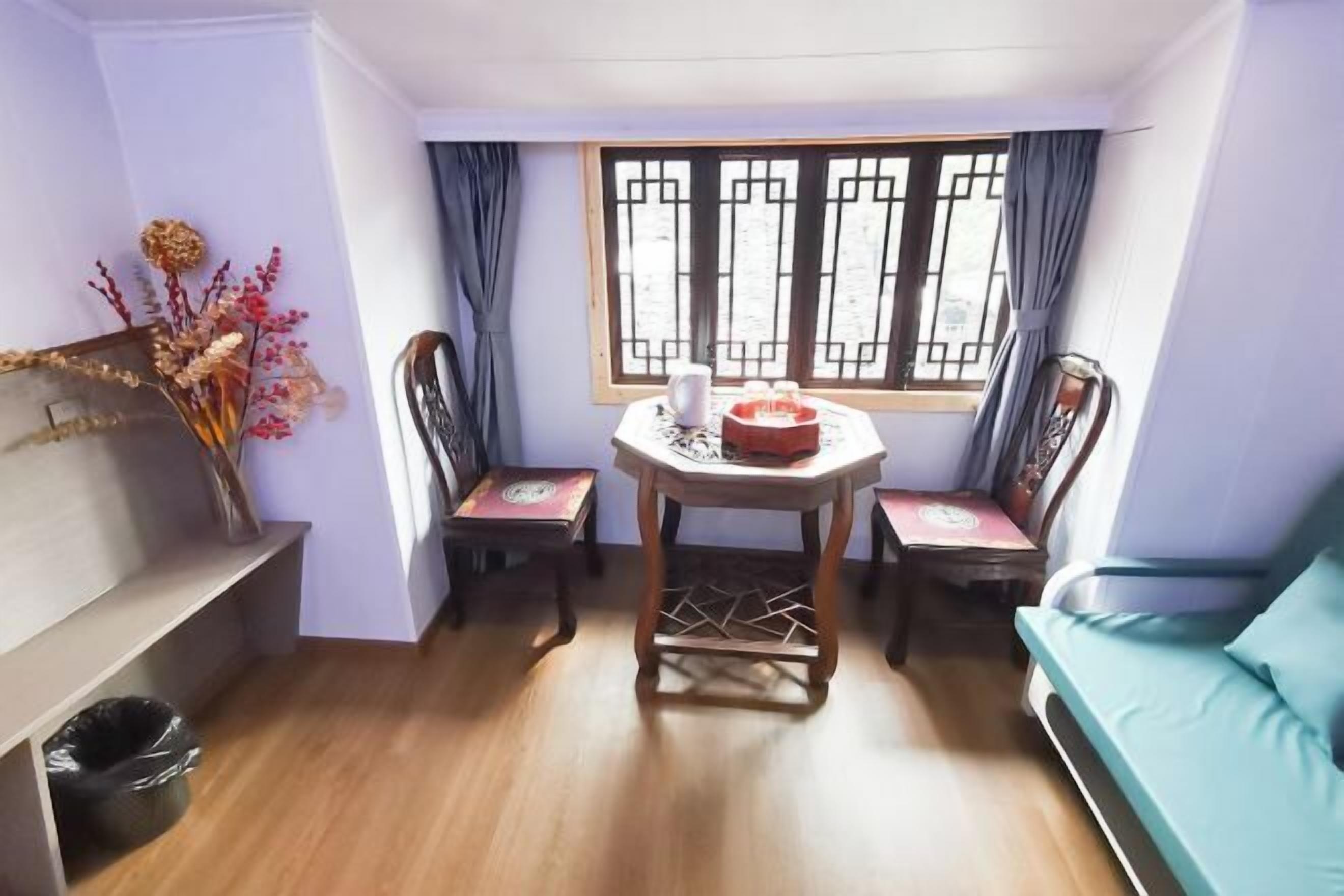 A room with a wooden floor, a table with chairs, a sofa, a window with curtains, and a vase with dried flowers.