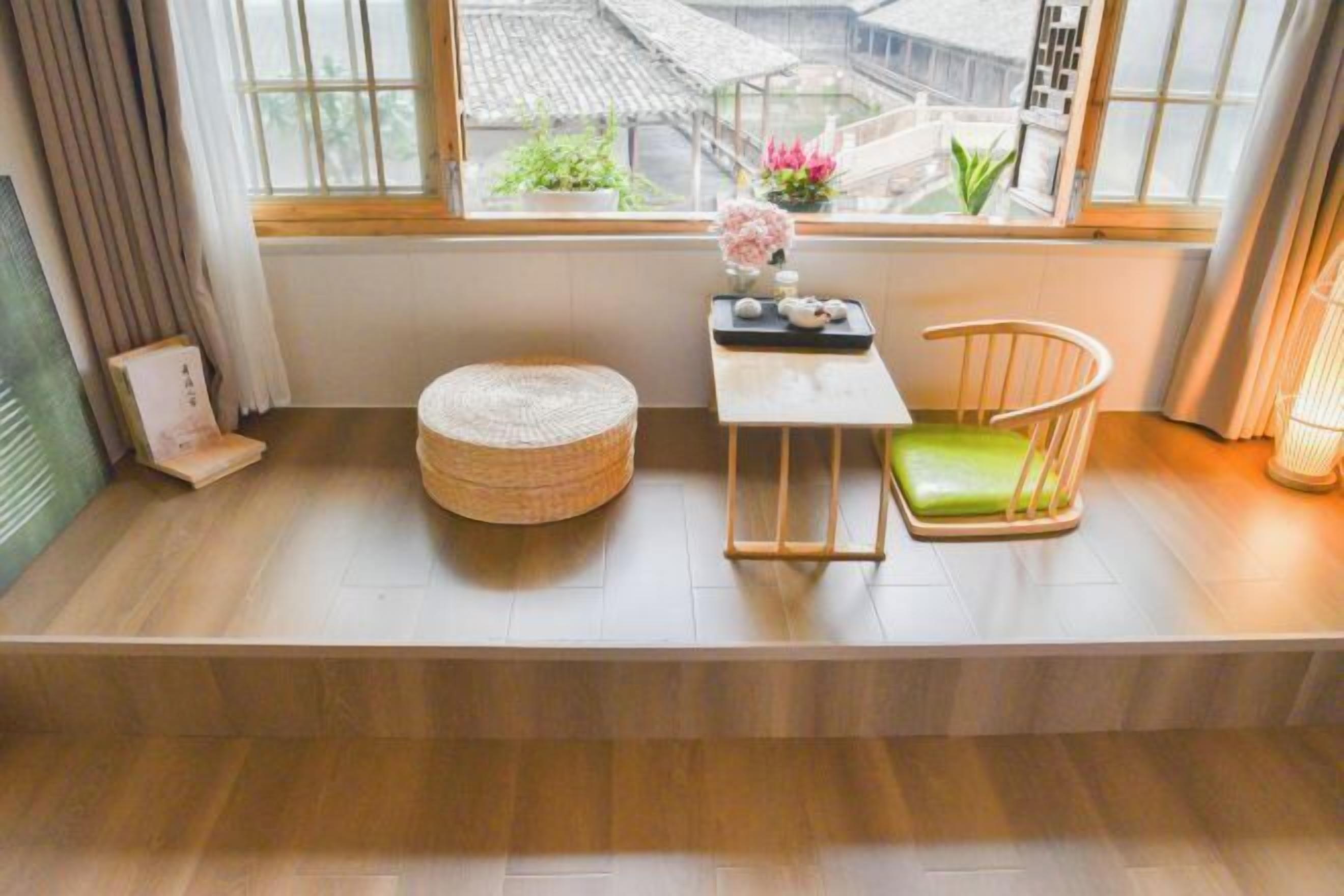 Una habitación con un banco de madera, una mesita con una bandeja, una silla con cojín verde y vistas a edificios a través de una ventana.