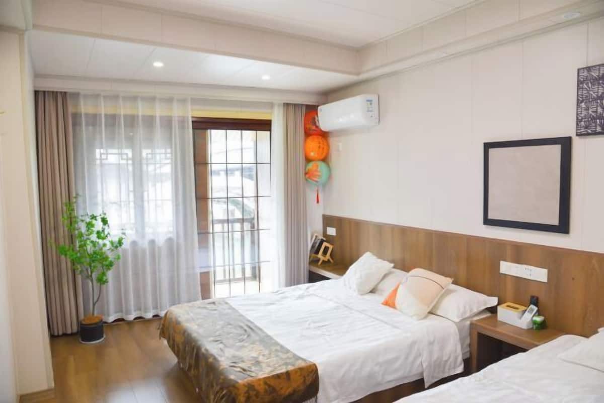 Hotellihuone, jossa on kaksi sänkyä, ruukussa oleva kasvi, ikkuna verhoilla, ilmastointilaite ja seinällä oleva kehystetty kuva.