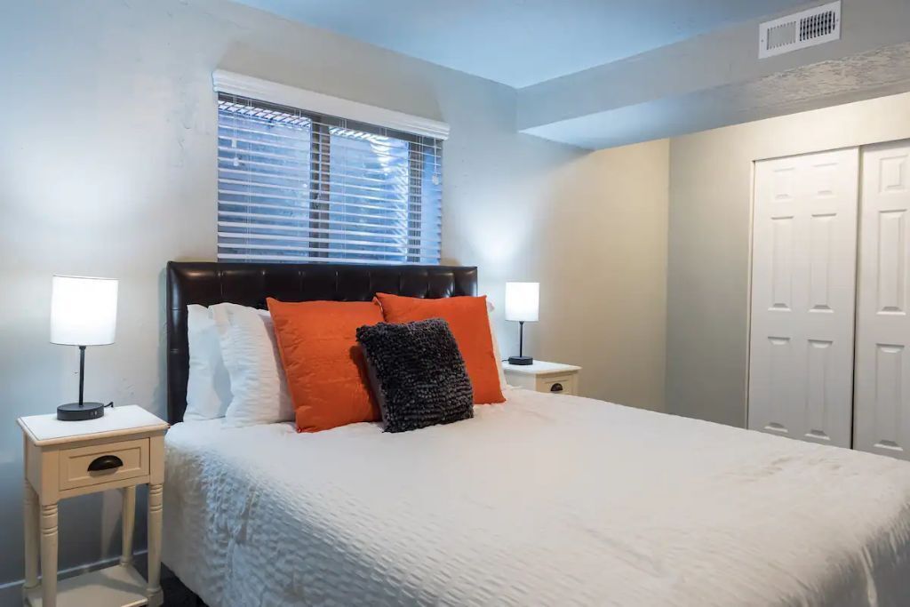 A bedroom with a bed, two bedside tables, a window with blinds, and a wardrobe.