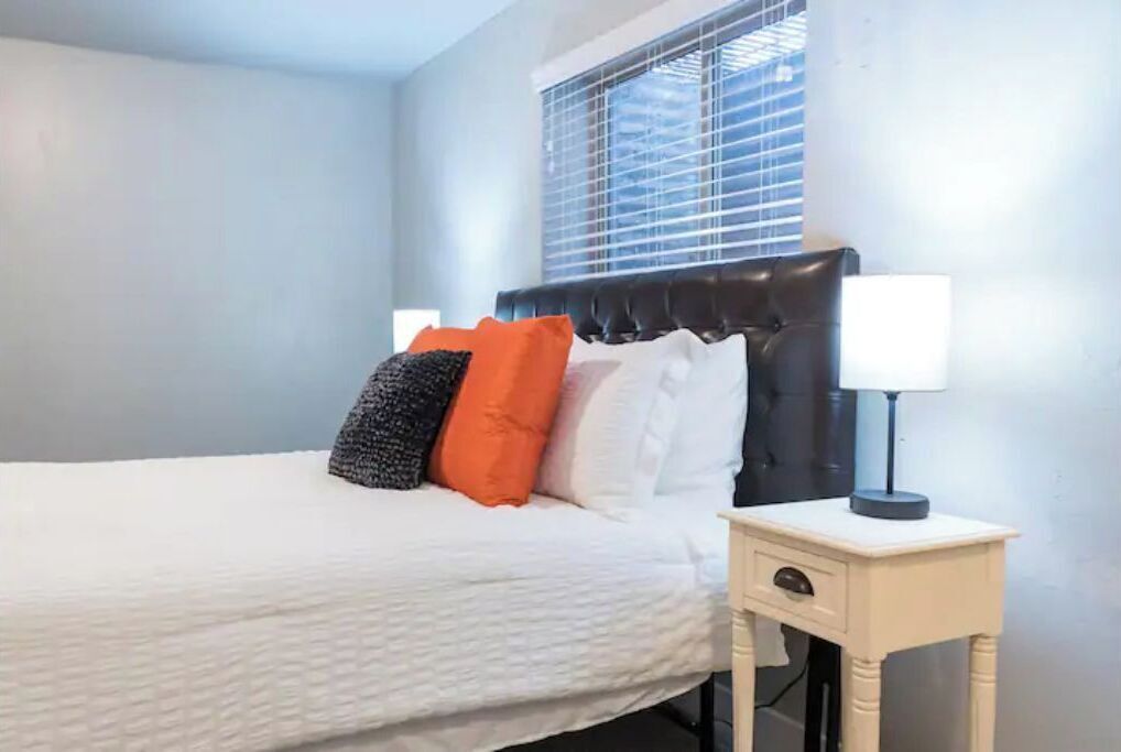 A neatly made bed with a white duvet, a black headboard, and several pillows in white, black, and orange. There is a bedside table with a lamp and a drawer.