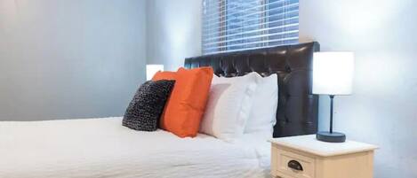 A neatly made bed with a white duvet, a black headboard, and several pillows in white, black, and orange. There is a bedside table with a lamp and a drawer.
