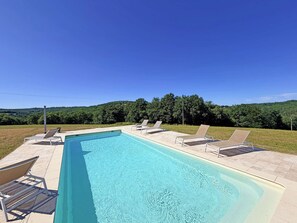 Una piscina rectangular con agua azul cristalina, rodeada por una terraza con baldosas. Cuatro sillas de descanso están alineadas a lo largo de la piscina. El fondo muestra un paisaje montañoso con árboles verdes y frondosos.