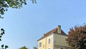 Un grand bâtiment à deux étages, doté d’une façade jaune et d’un toit recouvert de tuiles rouges, situé à proximité d’une étendue d’eau, laquelle est protégée par une barrière en béton et des poteaux.