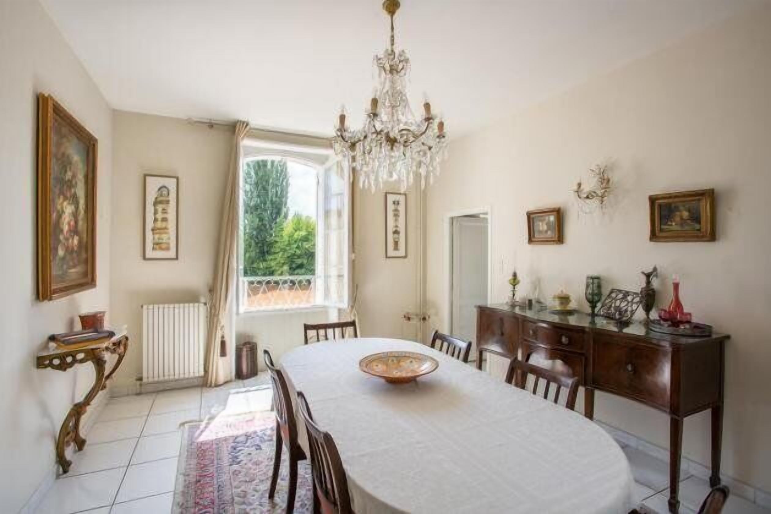 A dining room with a round table, chairs, a chandelier, and a view of greenery outside.