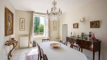 A dining room with a round table, chairs, a chandelier, and a view of greenery outside.