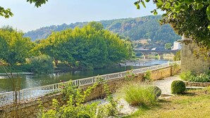 Une promenade au bord de la rivière, avec des marches en pierre et une rambarde blanche, entourée de verdure et offrant une vue sur un pont et des bâtiments au loin.