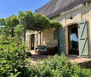 A stone building with a covered patio, a wooden bench, and a door with shutters. - Le Vezou (PAUNAT)