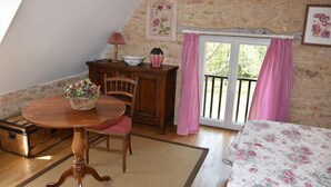 A cozy room with a stone wall, a wooden table with chairs, a bed with floral bedding, and a window with pink curtains.