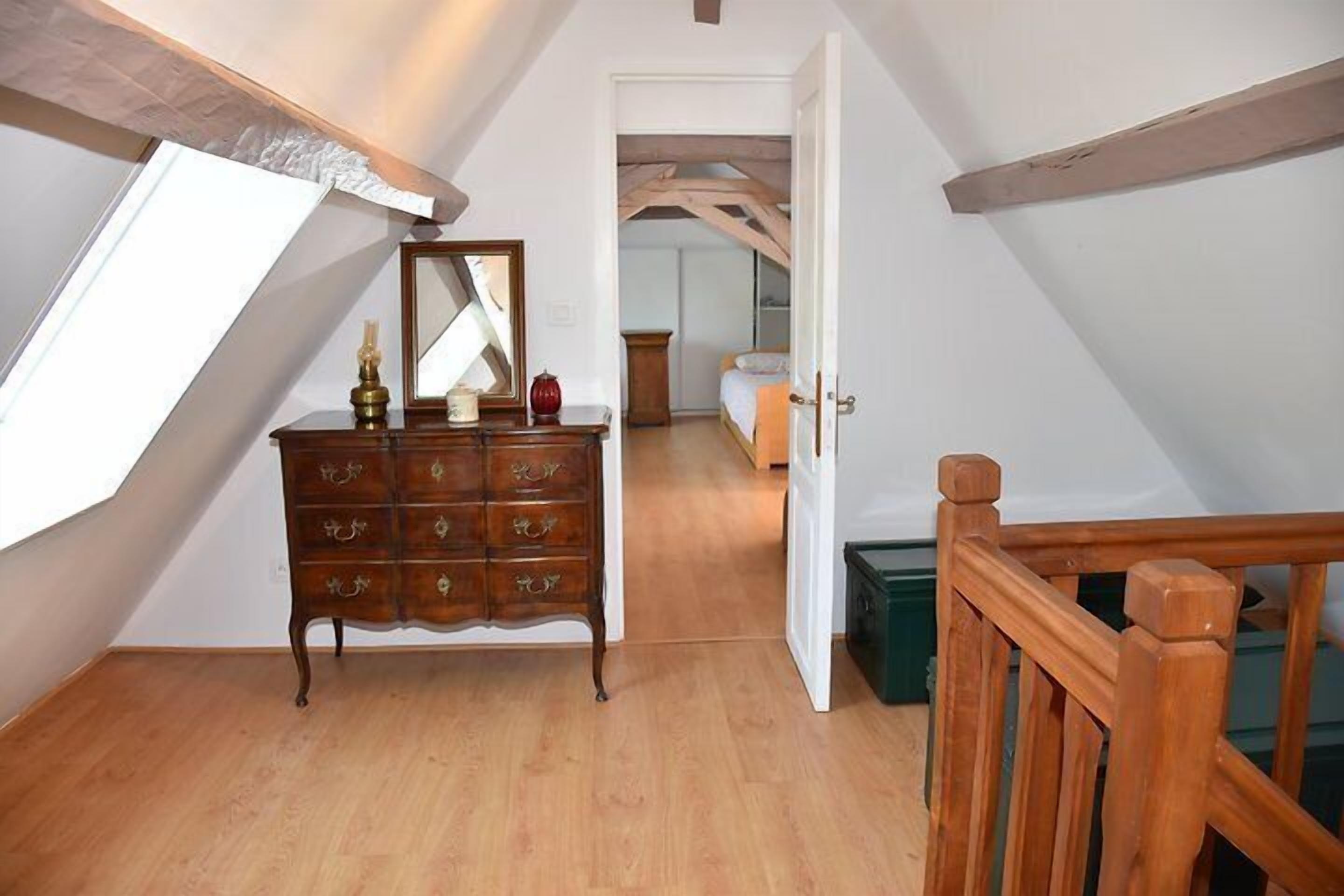 Une pièce avec une commode en bois, un miroir et une vue sur une chambre à coucher, visible à travers une porte ouverte.